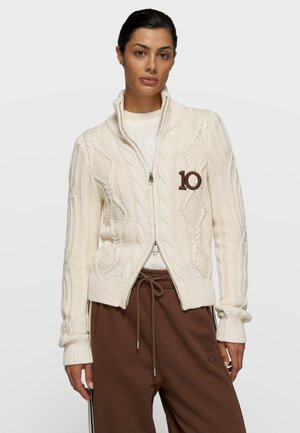 Kobieta ubrana w kremowy sweter na zamek z grubym splotem warkocza z naszywką "10", białą koszulę pod spodem oraz brązowe spodnie ze ściągaczem i bocznymi paskami.