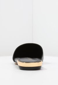 Svart slidesandal med en slät ovansida, guldaccentband och en platt sula. Insidan har en mjuk textur för komfort.