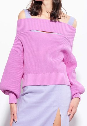 Femme portant un pull rose épaules dénudées sur une robe violet clair avec une fente haute sur la cuisse, debout devant un fond blanc.
