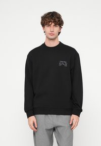 Μαύρο φούτερ με λαιμόκοψη τύπου crew, διακοσμημένο με λευκό λογότυπο στο αριστερό μέρος του στήθους. Το υλικό φαίνεται απαλό και με υφή, με ριμπ μανσέτες.