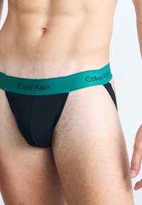 Schwarzer Stoffstring mit einem grünen elastischen Bund, auf dem "Calvin Klein" in schwarzer Schrift gedruckt ist. Glatte Textur, enger Sitz, minimalistisches Design.