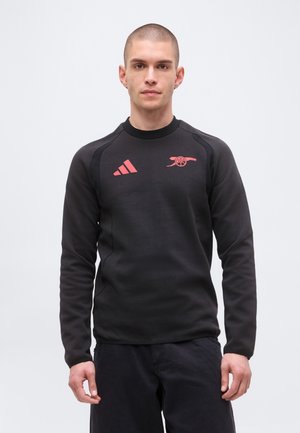 Ung mand iført en sort langærmet trøje med rød Adidas-logo og Arsenal-kanonemblem, stående mod en ensfarvet hvid baggrund.