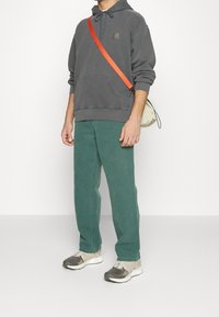 Persona che indossa un felpa grigia, pantaloni verdi, sneakers beige e grigie, e una borsa a tracolla bianca con una tracolla arancione brillante, su uno sfondo bianco.