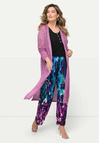 Larga chaqueta de punto ligera en color rosa sobre una blusa negra, combinada con pantalones sueltos de múltiples colores que presentan un atrevido patrón en púrpura, azul y verde.