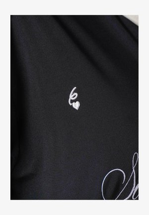 Tela negra con un pequeño diseño bordado en blanco de una mano haciendo un gesto de lengua de signos sobre una forma de corazón.