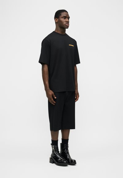 Άνδρας που στέκεται με μαύρο υπερμεγέθες T-shirt με μικρό κίτρινο κείμενο, μαύρα φαρδιά σορτς, μαύρες κάλτσες και λαμπερά μαύρα μποτάκια αστραγάλου, σε λευκό φόντο.