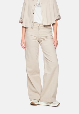 Frau trägt beige weit geschnittene Hosen, eine dazu passende kurze Jacke mit Schlangenmuster an den Manschetten, weiße Sneaker und ein weißes Oberteil mit Spitzen-Detail.