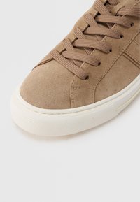 Ανοιχτό καφέ σουέντ αθλητικό παπούτσι με κορδόνια στο ίδιο χρώμα και λευκή υφή από καουτσούκ στη σόλα, γωνία που δείχνει τη μύτη και μέρος του πλάγιου μέρους.