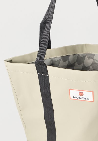 Saco tote impermeável bege com alças tecidas pretas, apresentando um forro geométrico cinza e uma etiqueta de marca com uma borda laranja.