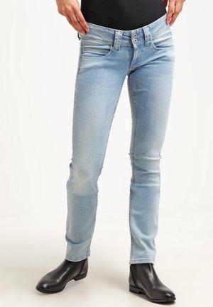 Persoon draagt lichtblauwe slim-fit jeans en zwarte enkellaarzen, houdt de tailleband vast om de losse pasvorm te tonen.