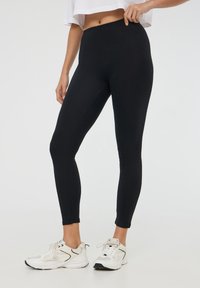 Leggings neri con una texture liscia, vita alta e design aderente, abbinate a scarpe da ginnastica bianche con un upper in rete e lacci.