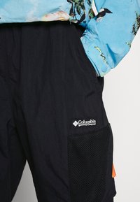 Närbild av svarta Columbia cargo-byxor med mesh-fickdetaljer och en person som bär en blå blommig skjorta med handen i fickan.