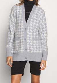 Cardigan grigio a motivo houndstooth con scollatura a V, chiusura con bottoni e due tasche frontali. Realizzato in morbido materiale a maglia con polsini a coste.