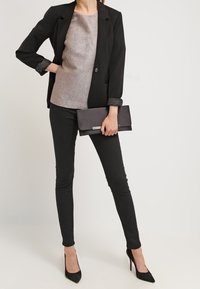 Žena v černém blejzru, kovovém topu, černých skinny kalhotách, držící černou kabelku a obutá v černých lodičkách, stojící na jednobarevném pozadí.