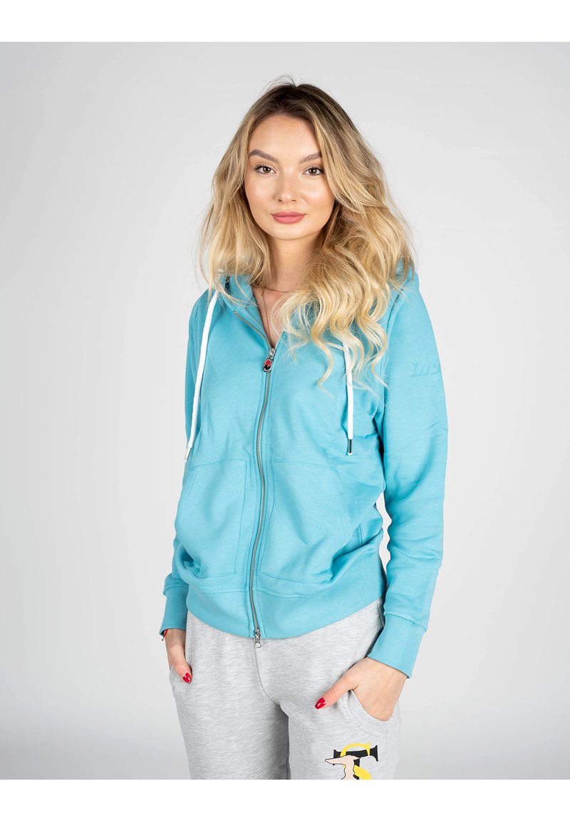 Felpa leggera blu con zip, dotata di tasca frontale e coulisse. Presenta una texture liscia e maniche aderenti, abbinata a pantaloni della tuta grigi.