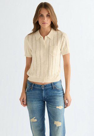 Beżowy krótki kardigan na drutach z krótkim rękawem, z pionowymi paskami, zapinany na guziki, z kieszenią na piersi, w połączeniu z przetartymi niebieskimi dżinsami.