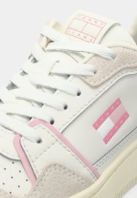 Close-up van een witte sneaker met roze accenten, veterdicht, met "Tommy Jeans" branding op de tong en het zijpaneel.