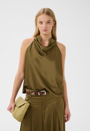 Junge Frau mit langen blonden Haaren trägt ein olivgrünes ärmelloses Oberteil und Hose und hält eine beige geflochtene Clutch vor einem schlichten Hintergrund.
