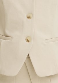 Blazer beige sur mesure avec deux boutons fermés et deux poches fendues horizontales à l'avant.
