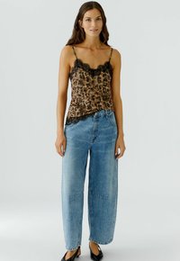 Luipaardprint cami-top met zwarte kantafwerking, gecombineerd met lichtgewassen wijde jeans. Model draagt zwarte ballerinashoes. Eenvoudige achtergrond.