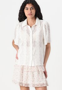 Blusa bianca in pizzo con maniche corte a palloncino e chiusura a bottoni, sovrapposta a una gonna trasparente a fantasia floreale in tonalità pastello con volant.