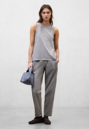 Femme portant un haut sans manches gris et un pantalon assorti, tenant un petit sac à main en tissu bleu, debout devant un fond gris uni.