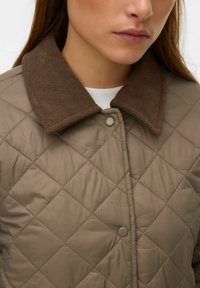 Veste matelassée beige avec un col en velours côtelé marron, dotée de boutons-pression et d'une texture en motif losange pour la chaleur et le style.
