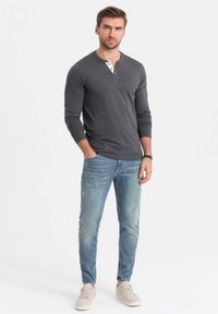 Mies seisoo kädet taskuissa tummanharmaassa pitkähihaisessa henley-paidassa, vaaleansinisissä farkuissa ja beigeissä tennareissa valkoista taustaa vasten.