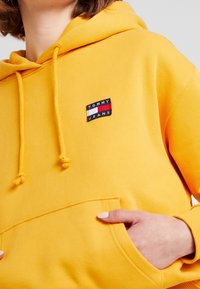 Sudadera amarilla con capucha y cordón, con un parche de marca en el pecho. Fabricada con tejido suave y con un bolsillo tipo canguro.