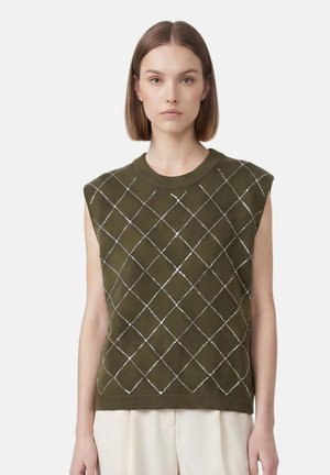 Jeune femme aux cheveux raides mi-longs, portant un pull sans manches vert olive avec un motif losange blanc et un pantalon beige.