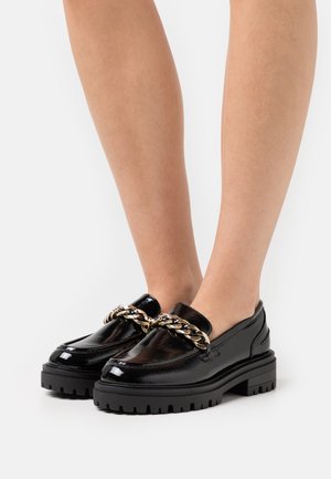 Musta lakattu nahka loafers paksulla mustalla pohjalla ja kultaketjua etuosassa. Suora pinta kiiltävällä viimeistelyllä.