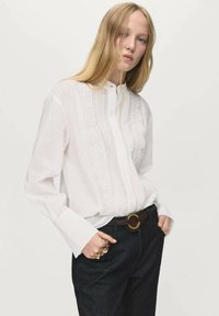 Witte blouse met lange mouwen met kantdetails en een knoopsluiting bij de kraag, gecombineerd met donkere spijkerbroek en een bruine riem met een ronde gesp.