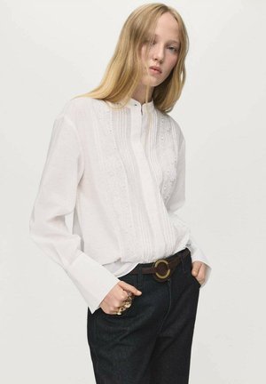 White long-sleeve blouse featuring lace detailing and a buttoned collar, paired with dark denim pants and a brown belt with a circular buckle.
