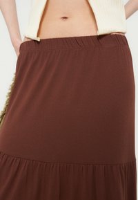 Femme portant une jupe marron taille haute avec une ceinture élastique et un top court côtelé crème, dévoilant le ventre et le nombril.