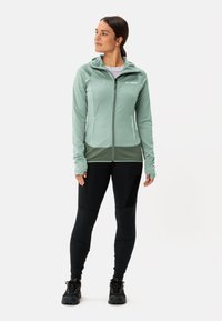 Giacca zip-up verde chiaro con finitura texturizzata, pannello inferiore verde scuro a contrasto e maniche con foro per il pollice. Indossata con leggings neri e scarpe.