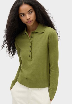Vrouw met lang krullend haar, gekleed in een olijfgroen poloshirt met lange mouwen en lichtgekleurde broek, kijkt naar links.
