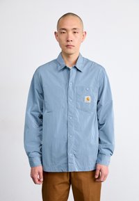 Chemise légère à manches longues, bleu, avec une poche poitrine et un patch logo ; texture lisse, coupe décontractée, fermeture par boutons.