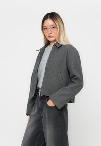 Giacca grigia corta con zip frontale, colletto con rever e tessuto testurizzato, abbinata a un maglione grigio chiaro e jeans scuri.