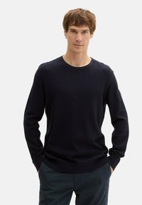 Dunkelblauer gerippter Pullover mit langen Ärmeln und rundem Halsausschnitt. Mit einem Logo-Patch am linken Ärmel und einem taillierten unteren Saum.