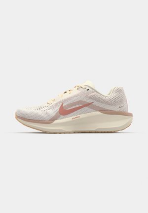 Chaussure de course tricotée beige clair avec le swoosh rose de Nike et une semelle amortie sur un fond gris clair uni.