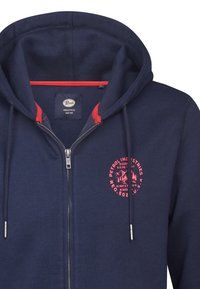 Felpa con zip di colore blu navy, con un accento rosso attorno al collo, caratterizzata da un logo circolare rosso sul petto e una cerniera in metallo argentato.