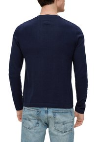 Granatowy, długi sweter dzianinowy z okrągłym dekoltem, gładką fakturą i ściągaczem na dole, w zestawieniu z jasnoniebieskimi dżinsami.