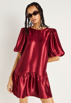 Robe en satin rouge avec une coupe ample, des manches bouffantes courtes et un ourlet en volants. Le tissu présente un éclat brillant et une texture lisse.