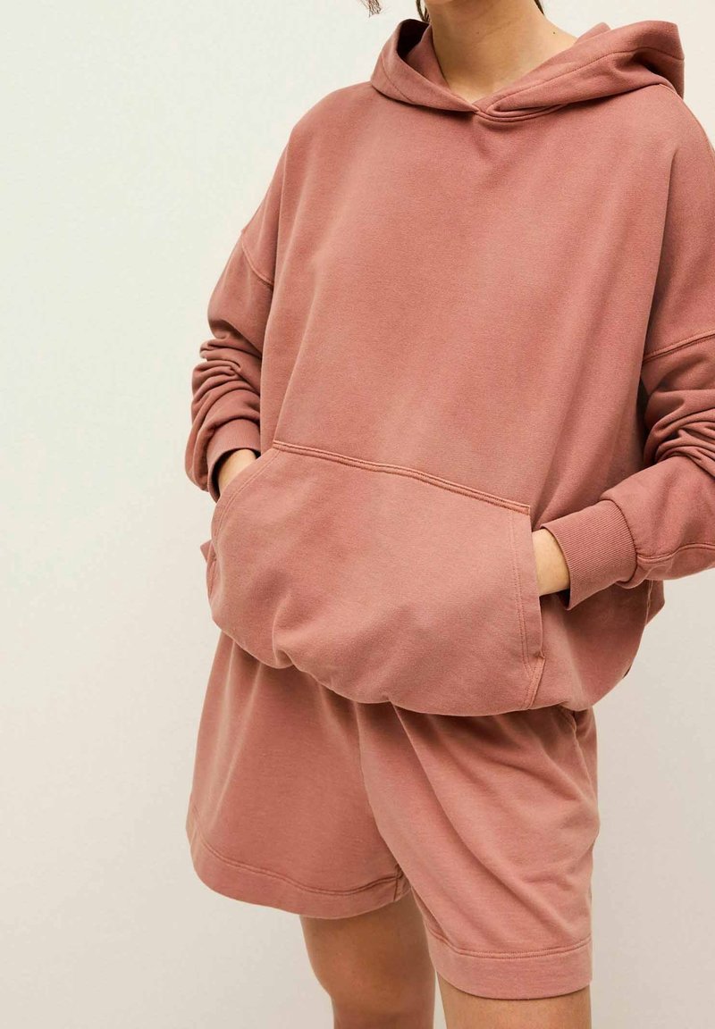 Sweat à capuche surdimensionné rose poudré avec une poche kangourou et des poignets côtelés, associé à un short assorti à coupe décontractée. Tissu doux en mélange de coton.