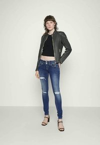 Črna skrajšana majica, raztrgane modre skinny jeans hlače in temno siva usnjena jakna z zadrgami. Model nosi črne sandale na peto.