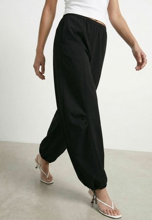 Femme marchant portant un pantalon de jogging noir à cordon, un haut blanc sans manches et des sandales blanches à talons avec brides sur un sol clair et un arrière-plan.