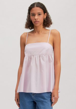 Femme aux cheveux bouclés portant un haut sans manches rose clair avec de fines bretelles et un jean bleu sur un fond uni.