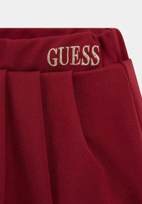 Pantaloni in tessuto rosso con design plissettato e vita elastica, con "GUESS" ricamato in oro. Texture liscia, stile casual.