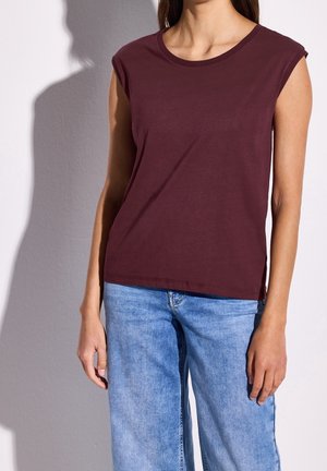 Kvinde iført vinrød ærmeløs top og lyseblå jeans, stående mod en hvid væg med synlig skygge.