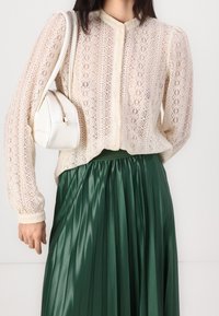 Lace blouse in creamy beige with a high neck and long sleeves, paired with a pleated green satin skirt and a small white rounded bag.